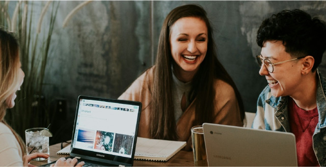 Three people sit at a table, laughing with each other as they work on laptops